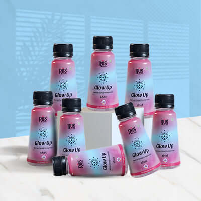 Eight bottles of Rus Organic's "Glow Up" wellness shot are arranged on and around a white platform with a blue background. The vibrant pink-to-blue gradient bottles feature a glowing radiance icon and black caps. Each 40ml shot is infused with amla, rose, and pomegranate – a nourishing blend designed to promote skin hydration, collagen production, and natural glow. Packed with antioxidants and Vitamin C, "Glow Up" supports clear, radiant skin from within.