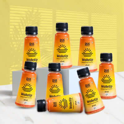 A vibrant 125ml bottle of Rus Organic's "WakeUp" shot is displayed on a wooden board surrounded by fresh ginger root, turmeric powder, and halved oranges. The orange-yellow gradient bottle features bold sunrise graphics, representing an energizing start to the day. Infused with orange, ginger, and turmeric, this wellness shot is designed to strengthen immunity, improve digestion, and support hydration with natural ingredients and antioxidants.