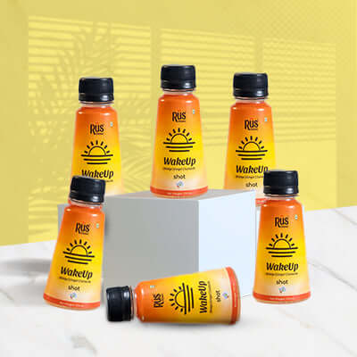 A vibrant 125ml bottle of Rus Organic's "WakeUp" shot is displayed on a wooden board surrounded by fresh ginger root, turmeric powder, and halved oranges. The orange-yellow gradient bottle features bold sunrise graphics, representing an energizing start to the day. Infused with orange, ginger, and turmeric, this wellness shot is designed to strengthen immunity, improve digestion, and support hydration with natural ingredients and antioxidants.