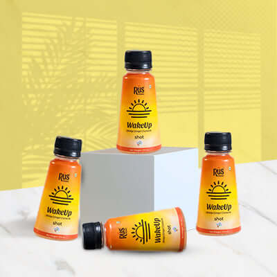 A vibrant 125ml bottle of Rus Organic's "WakeUp" shot is displayed on a wooden board surrounded by fresh ginger root, turmeric powder, and halved oranges. The orange-yellow gradient bottle features bold sunrise graphics, representing an energizing start to the day. Infused with orange, ginger, and turmeric, this wellness shot is designed to strengthen immunity, improve digestion, and support hydration with natural ingredients and antioxidants.